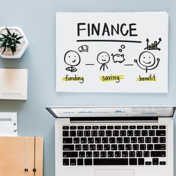 Stůl na němž je papír s nápisem FINANCE a pod ním jásající postavičky a jedna z nich získala benefit, na stole je dále počítač, diář a květina.