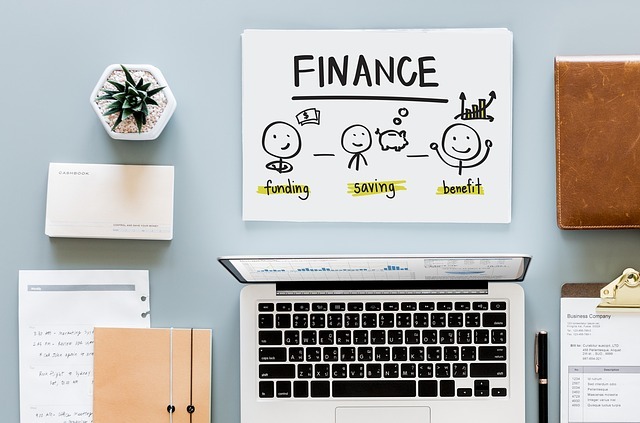 Stůl na němž je papír s nápisem FINANCE a pod ním jásající postavičky a jedna z nich získala benefit, na stole je dále počítač, diář a květina.